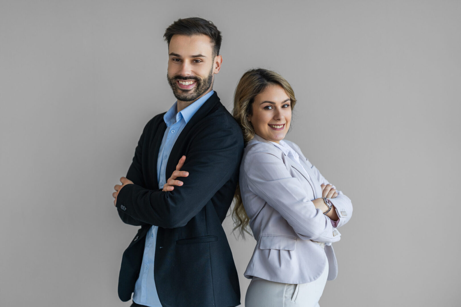 A confident man and woman in smart blazers stand back-to-back, smiling in a neutral studio setting. The scene conveys teamwork, professionalism, friendship, and modern business style.
