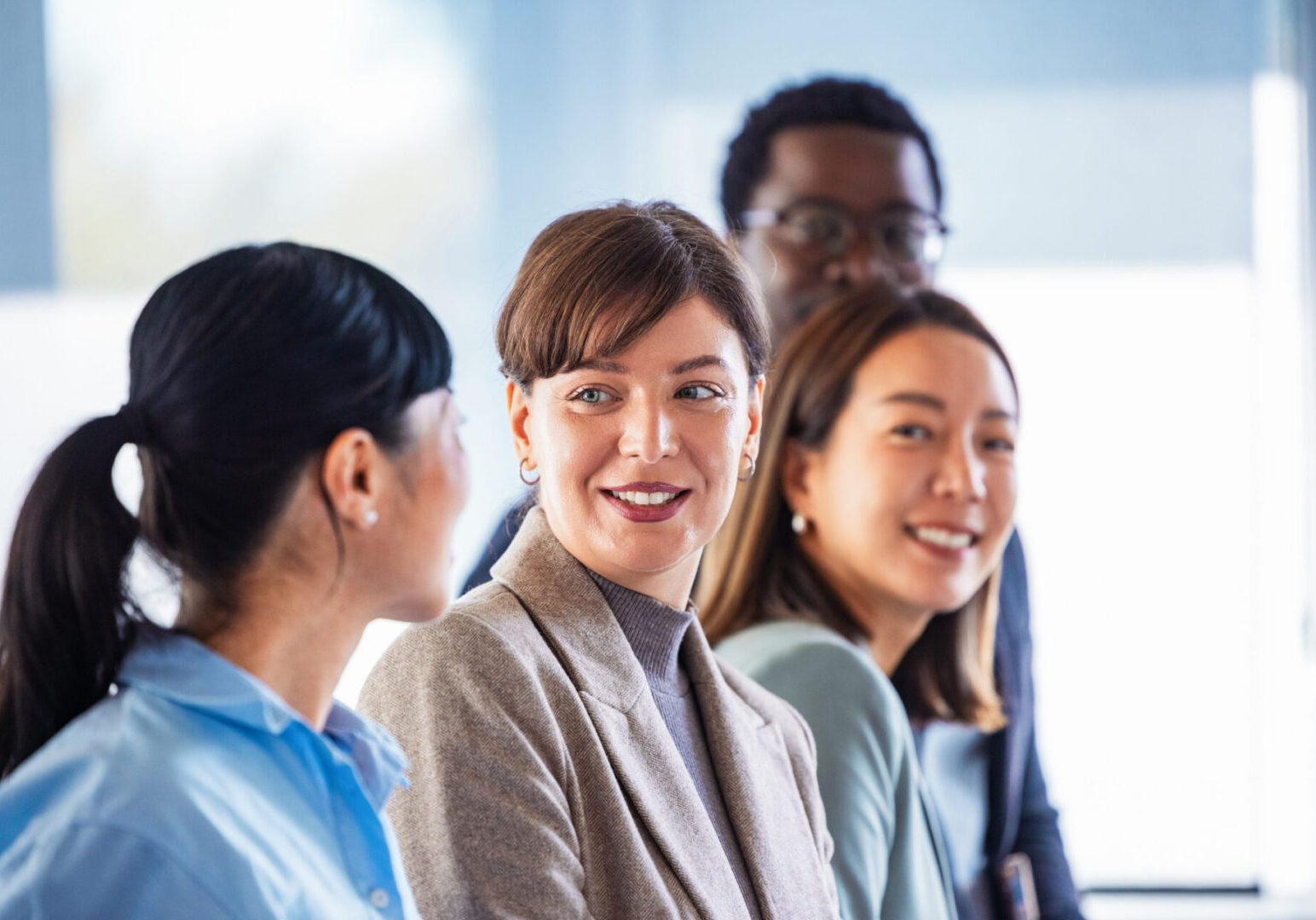 A diverse group of coworkers in a bright office engages in a relaxed discussion. A central smiling woman leads the moment while others listen, signaling teamwork, inclusivity, and a positive business vibe.
